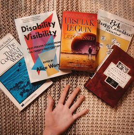 Five paperbacks, one of each of my five fingers pointing to a book. Left to right: The Three-Cornered World by Soseki, Disability Visibility by Wong, The Dispossessed by Le Guin, 84 Charing Cross Road by Hanff, Oranges Are Not the Only Fruit by Winterson.