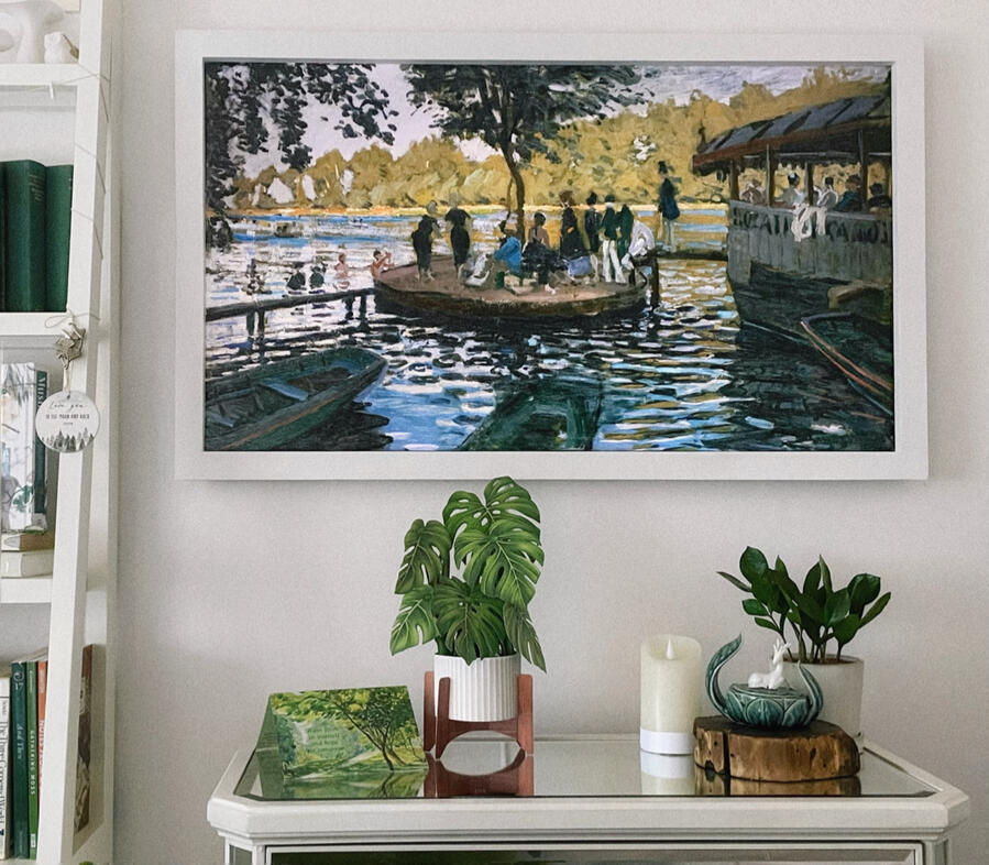 A white-framed TV hangs over a mirrored dresser. The TV shows the painting La Grenouillère by Monet. On the dresser are a green card, paper monstera plant, faux candle, sculpture of a deer on a lotus, and ZZ plant. A bit of bookshelf seen on the left.