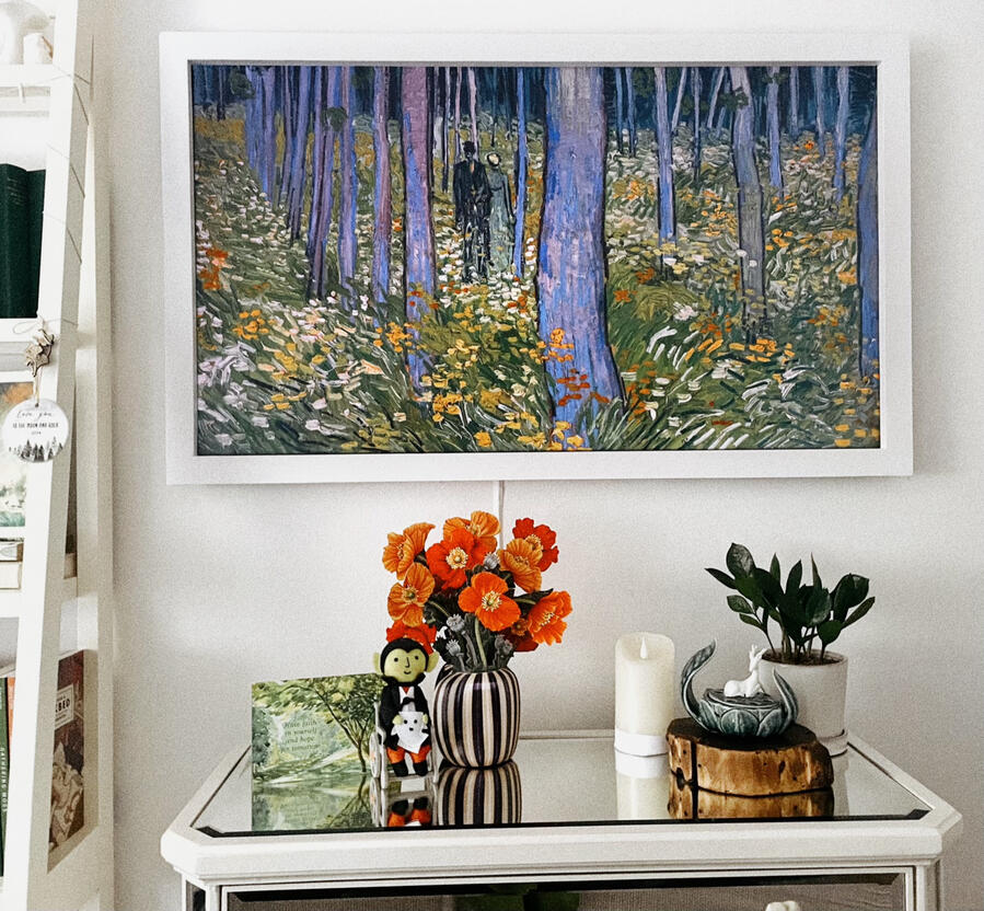 View from My Bed A white-framed TV hangs over a dresser. The TV shows the painting Undergrowth With Two Figures by Van Gogh. On the dresser are a green card, felt Dracula in a wheelchair, paper orange poppies, faux candle, sculpture of a deer on a lotus, and ZZ plant.
