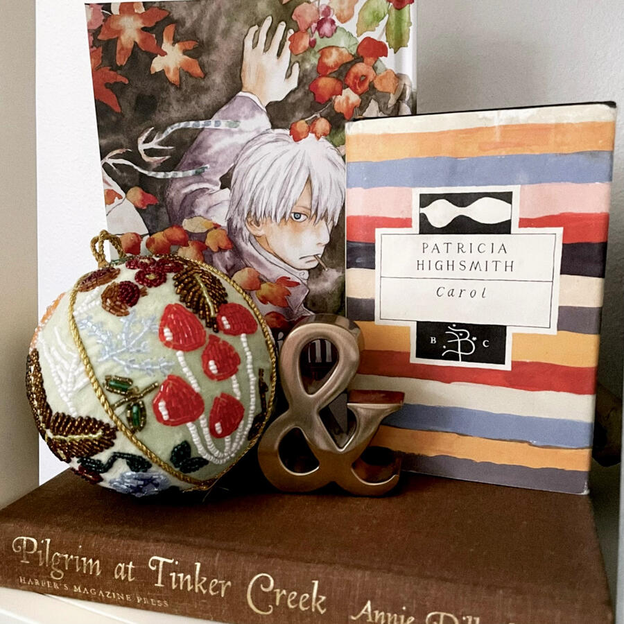 A Bookshelf Vignette Three hardcover books are displayed together with beaded holiday ornament and a golden ampersand paperweight. The look is autumnal, orange, red, and brown. Fall leaves and mushrooms. The books are Mushishi, Carol, and Pilgrim at Tinker Creek.