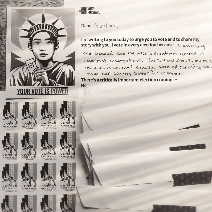 Black-and-white. A sticker showing a young person wearing the crown of the Statue of Liberty says,”Your vote is power”. Nearby is a hand-written letter to a voter, a stack of envelopes, and stamps with Women’s Suffrage designs.